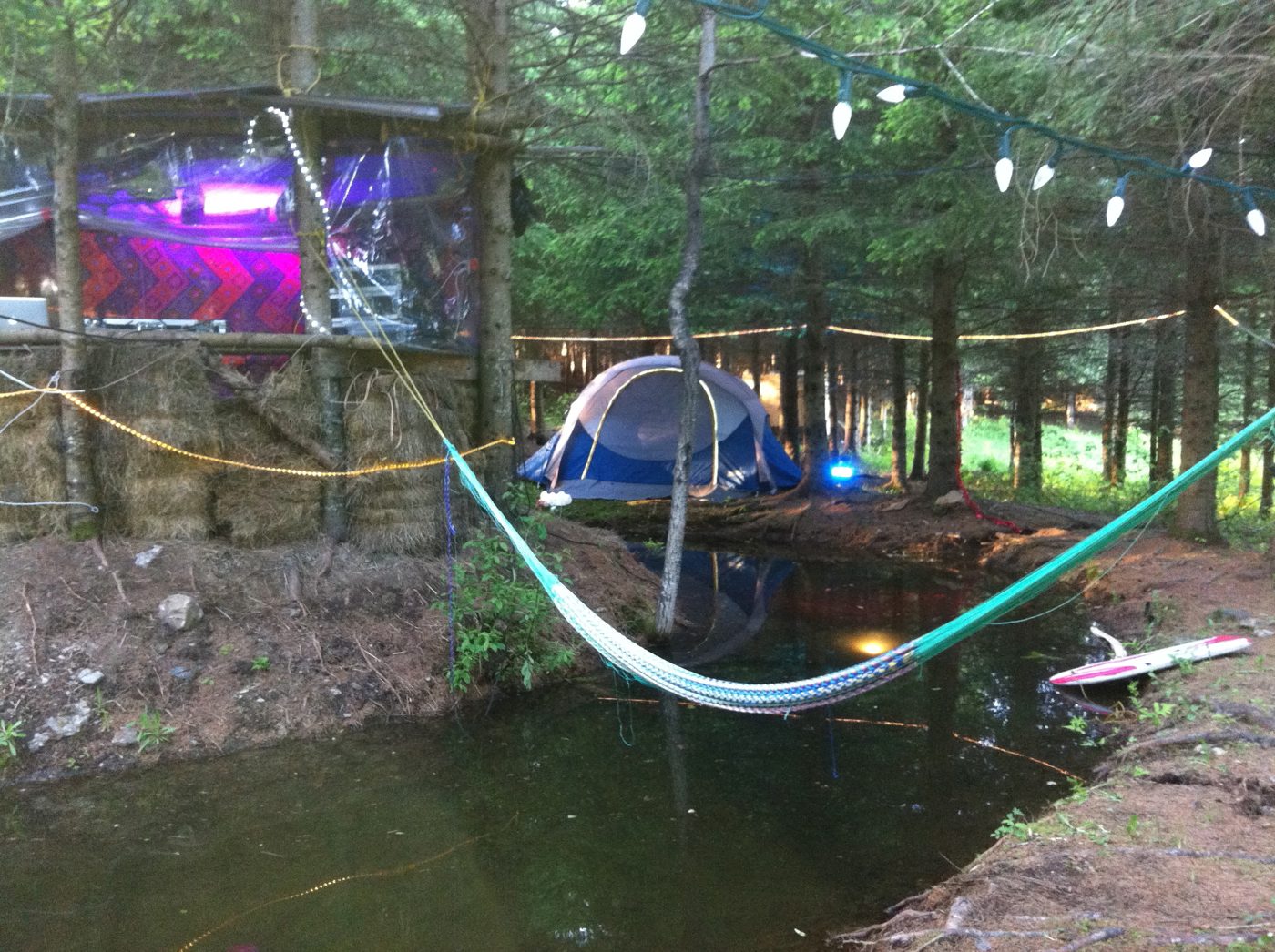 Le Cherry Island de L'AgorA : la cabane au bord de l'eau, le hamac tendu au-dessus du ruisseau, une tente sous les épinettes, des guirlandes entre les arbres.